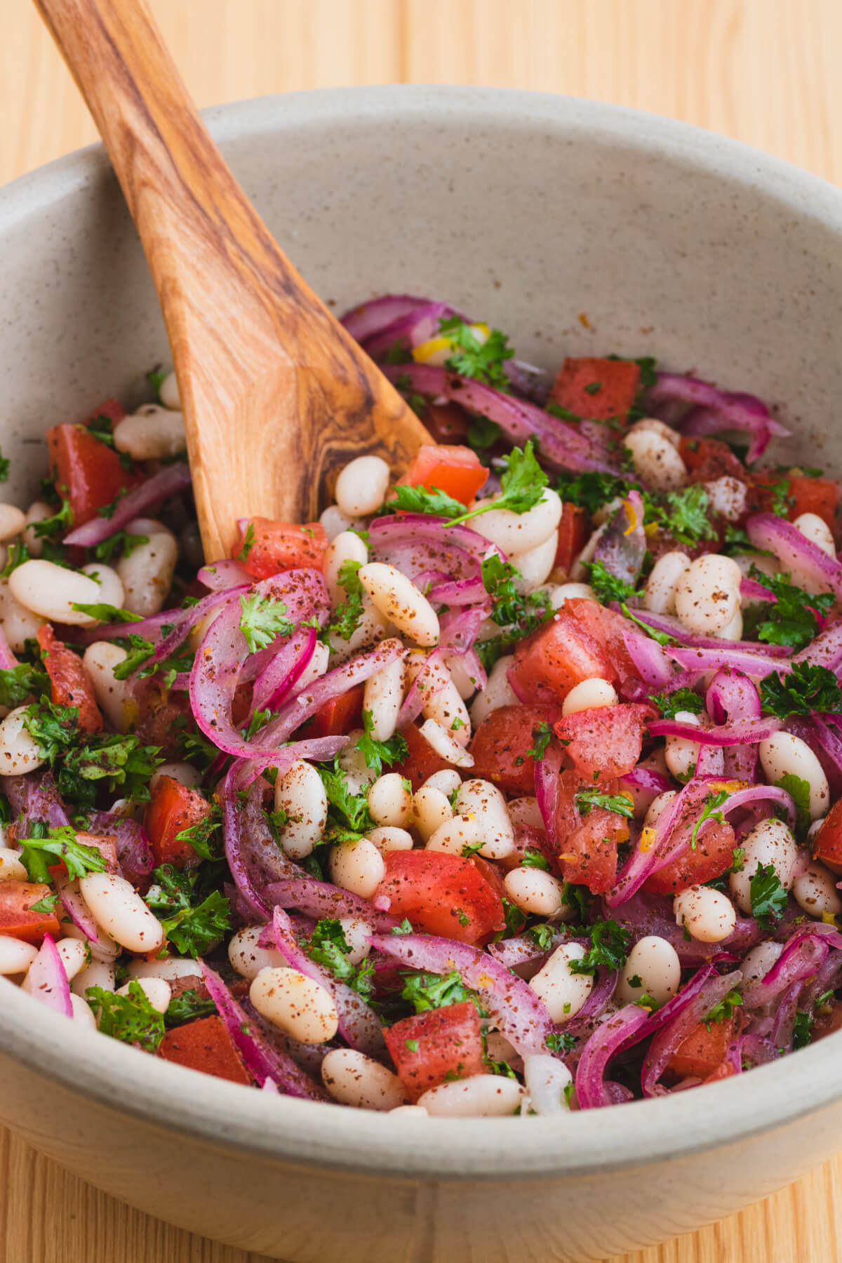 A wooden spoon holds a serving of colourful white bean salad with red onions, tomatoes, and green curly parsley.