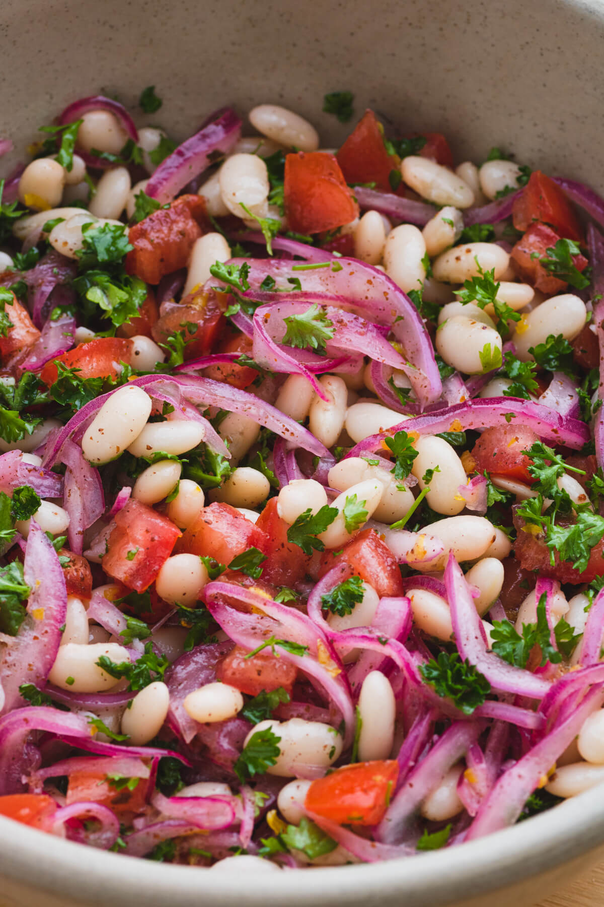 A bowl full of colourful white bean salad with red onions, tomatoes, and green curly parsley.