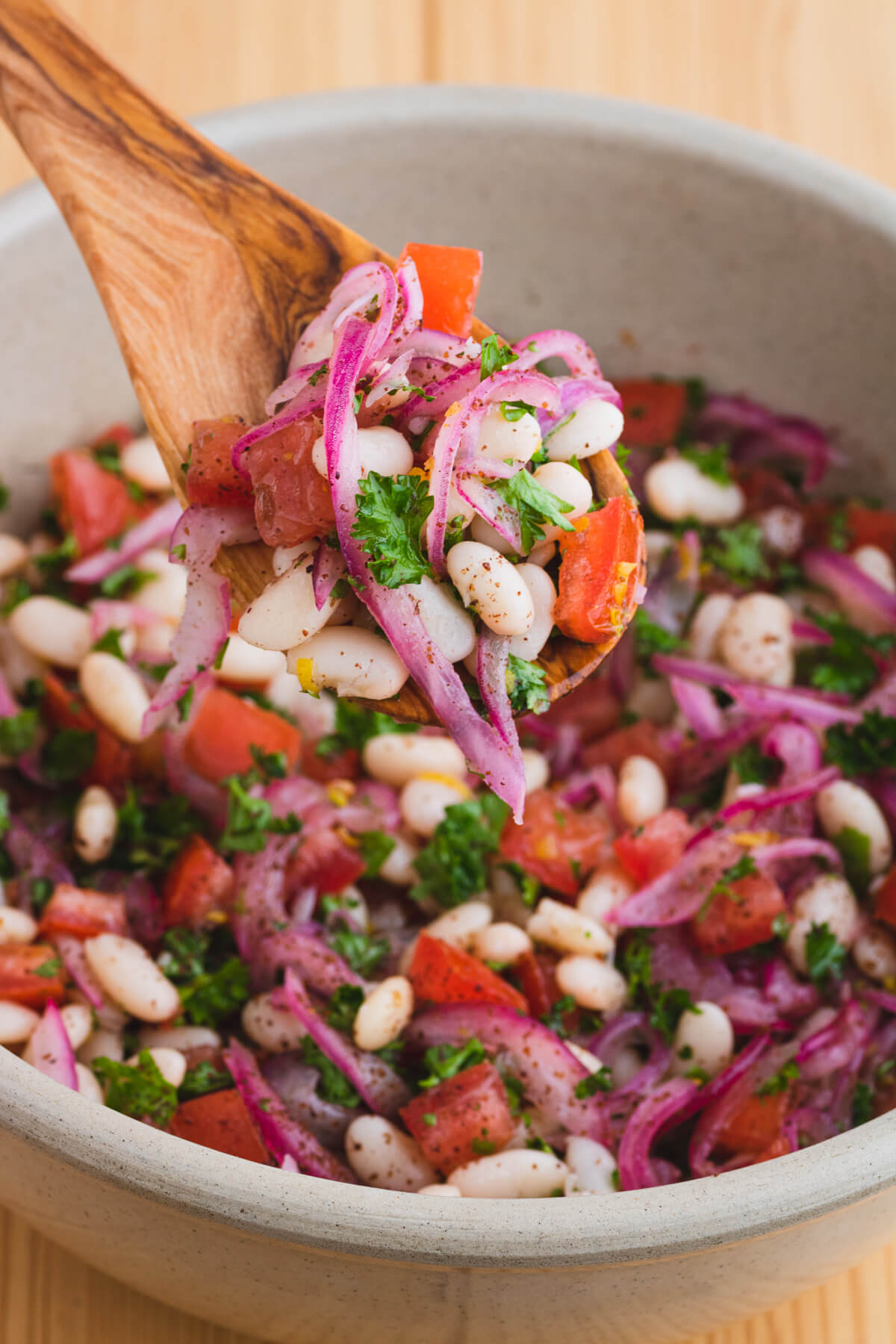 A wooden spoon holds a serving of colourful white bean salad with red onions, tomatoes, and green curly parsley.