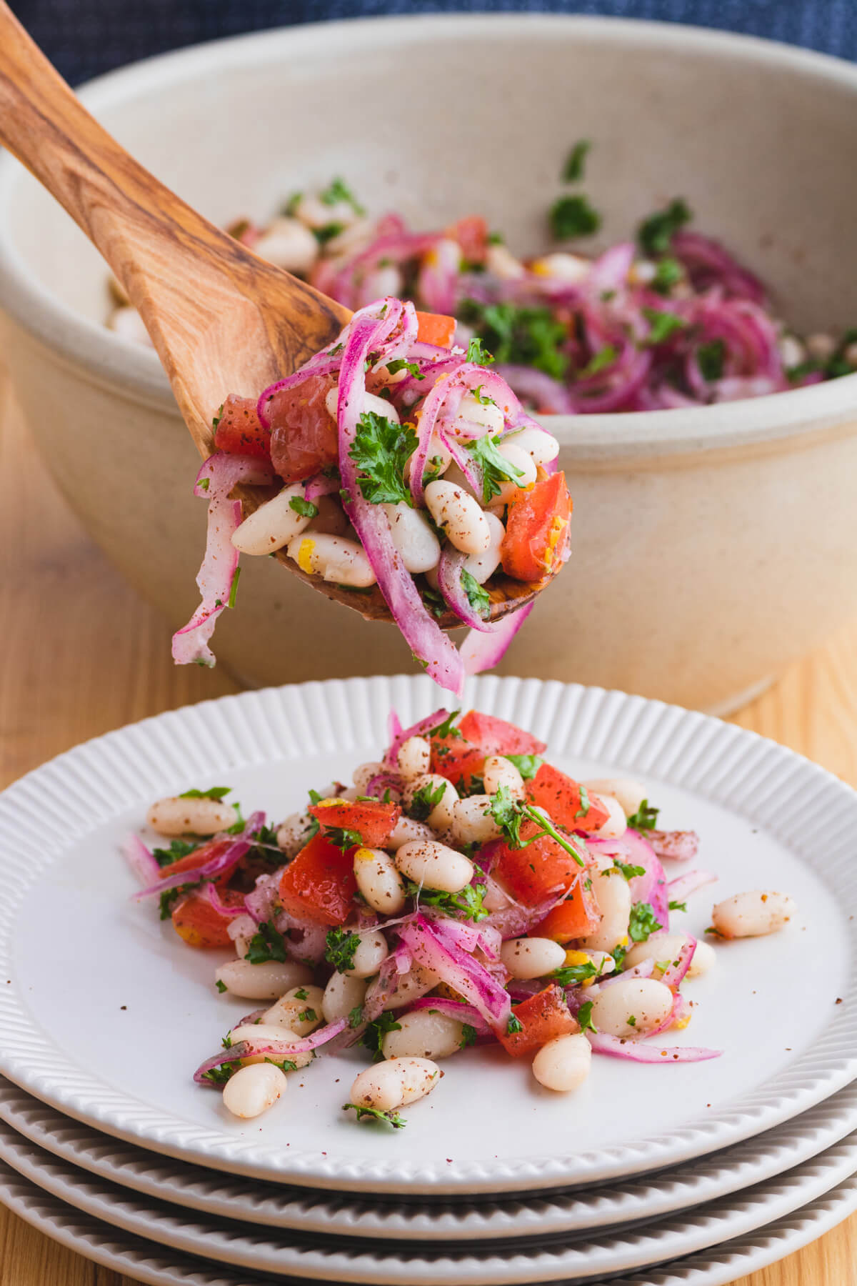 A wooden spoon serves a colourful white bean salad with red onions, tomatoes, and green curly parsley onto a white plate.