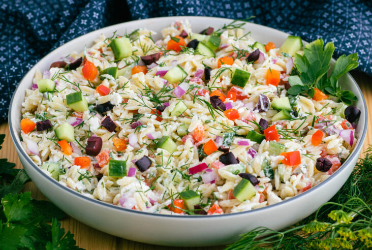 A white bowl holding a colourful Greek Orzo salad with feta cheese, green herbs, cucumbers, tomatoes, black olives, and peppers.