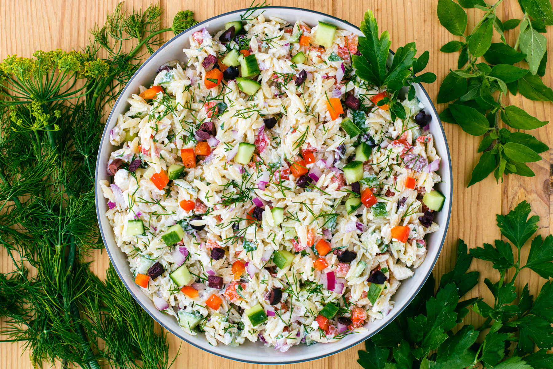 A white bowl holding a colourful Greek Orzo salad with feta cheese, green herbs, cucumbers, tomatoes, black olives, and peppers on a wooden background.