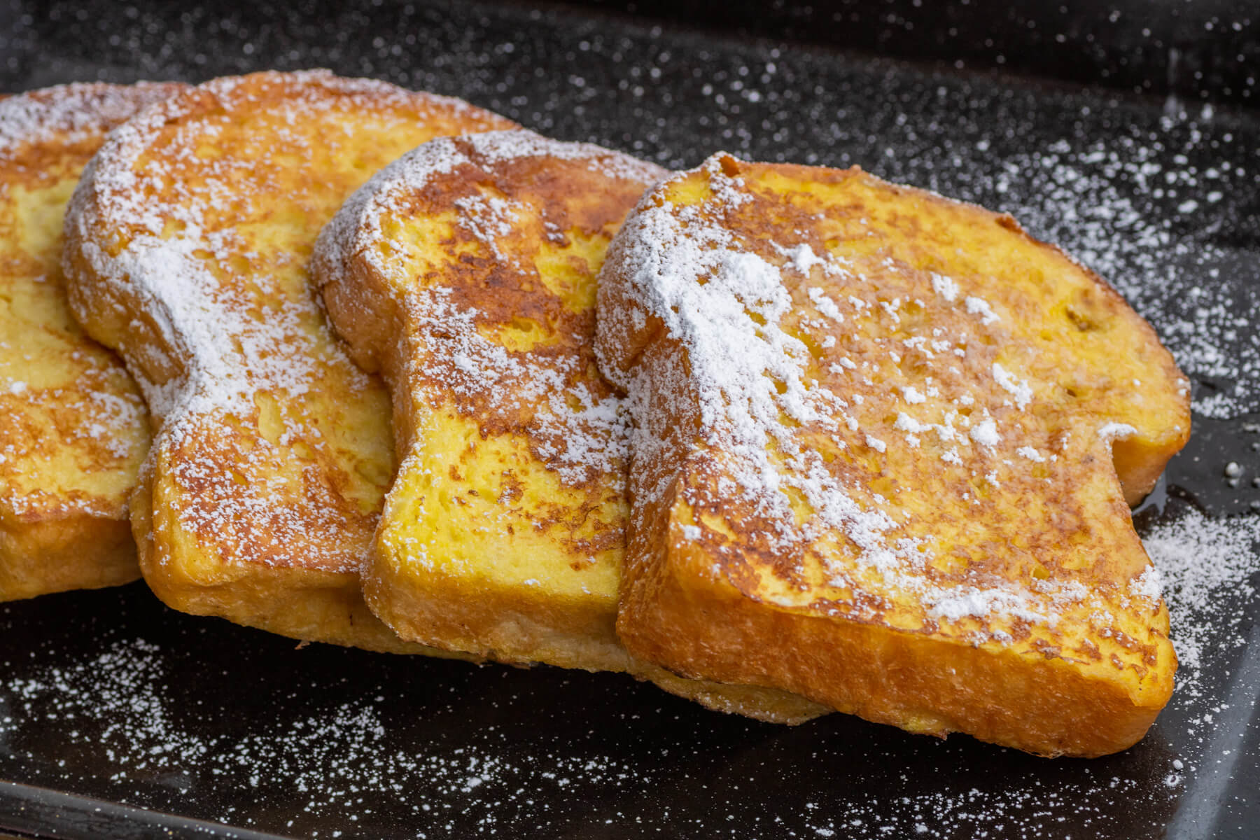A black tray containing slices of golden Brioche French toast dusted with confectioner's sugar.