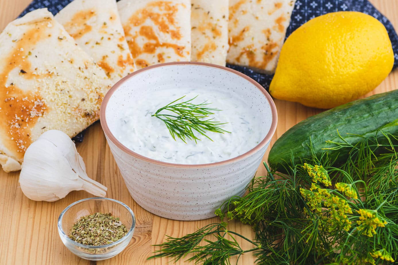 A small bowl of creamy white Tzatziki sauce flecked with cucumber and dill surrounded by grilled pita bread and other ingredients.