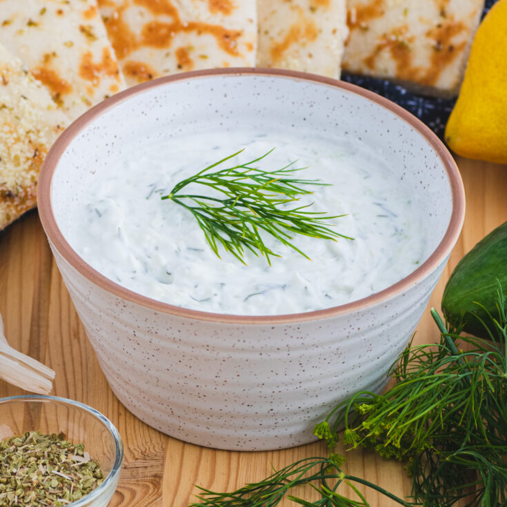 A small bowl of creamy white Tzatziki sauce flecked with cucumber and dill surrounded by grilled pita bread and other ingredients.