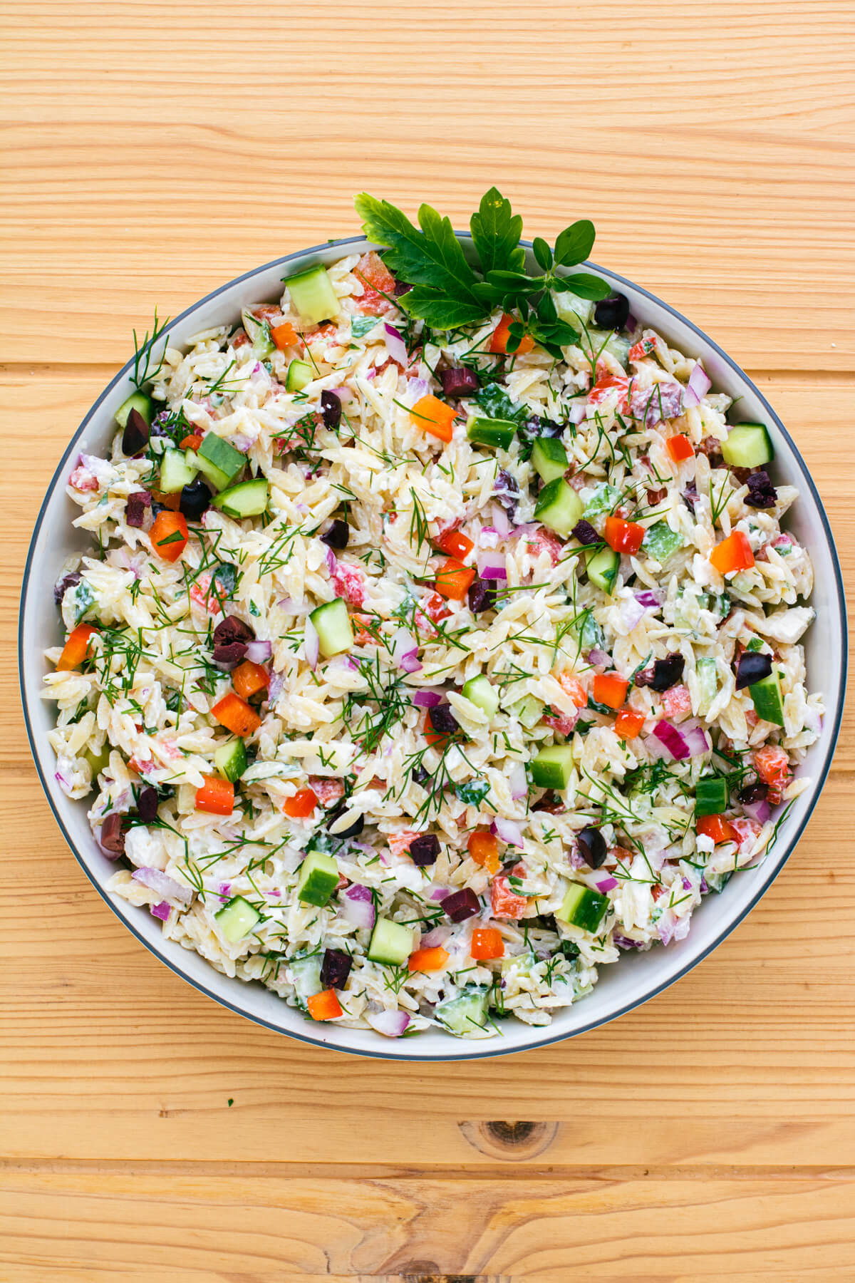 A white bowl holding a colourful Greek Orzo salad with feta cheese, green herbs, cucumbers, tomatoes, black olives, and peppers on a wooden background.