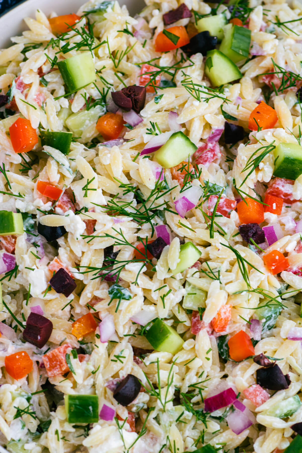 Close up of a colourful Greek Orzo salad with feta cheese, green herbs, cucumbers, tomatoes, black olives, and peppers.