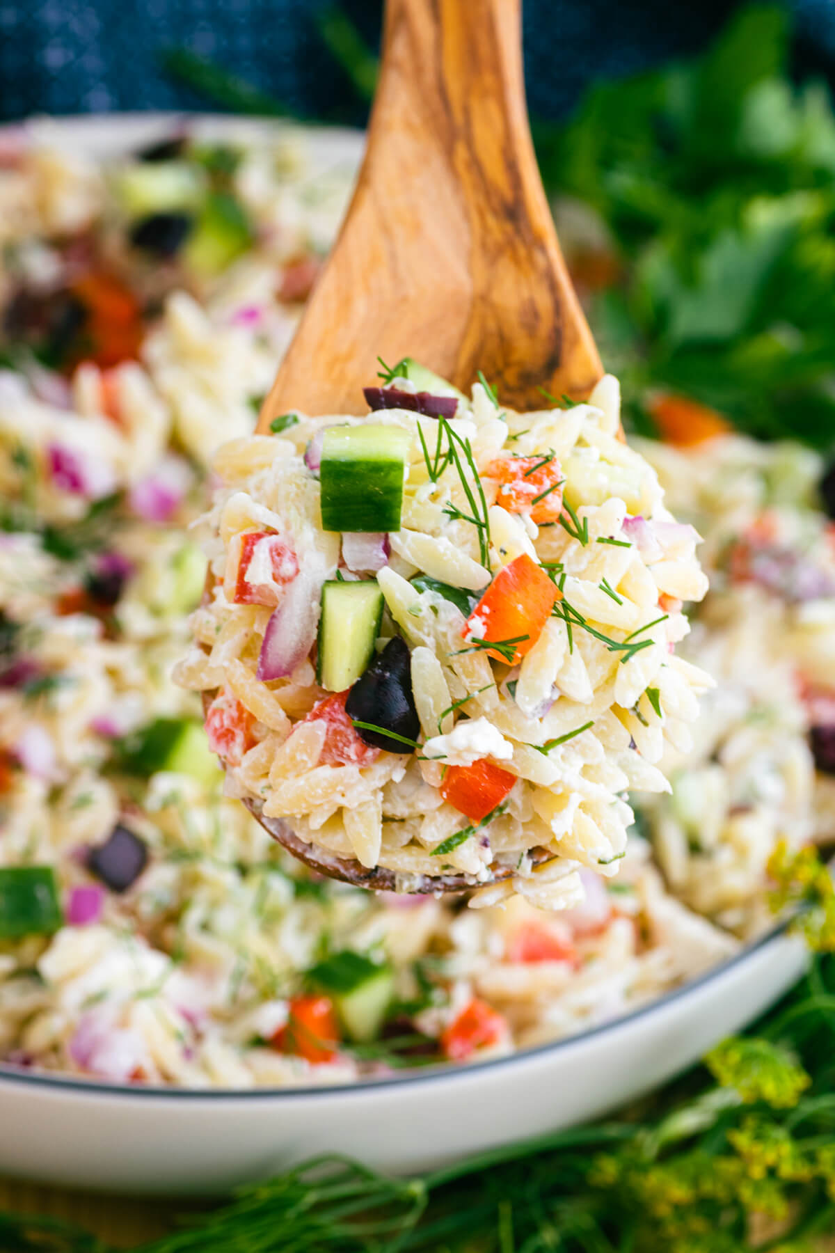 A wooden spoon full of holding colourful Greek Orzo salad with feta cheese, green herbs, cucumbers, tomatoes, black olives, red onions, and peppers.