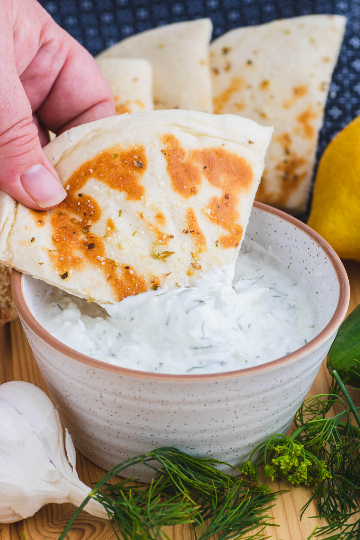A hand dips a toasted triangle of pita bread in a bowl of creamy tzatziki sauce.