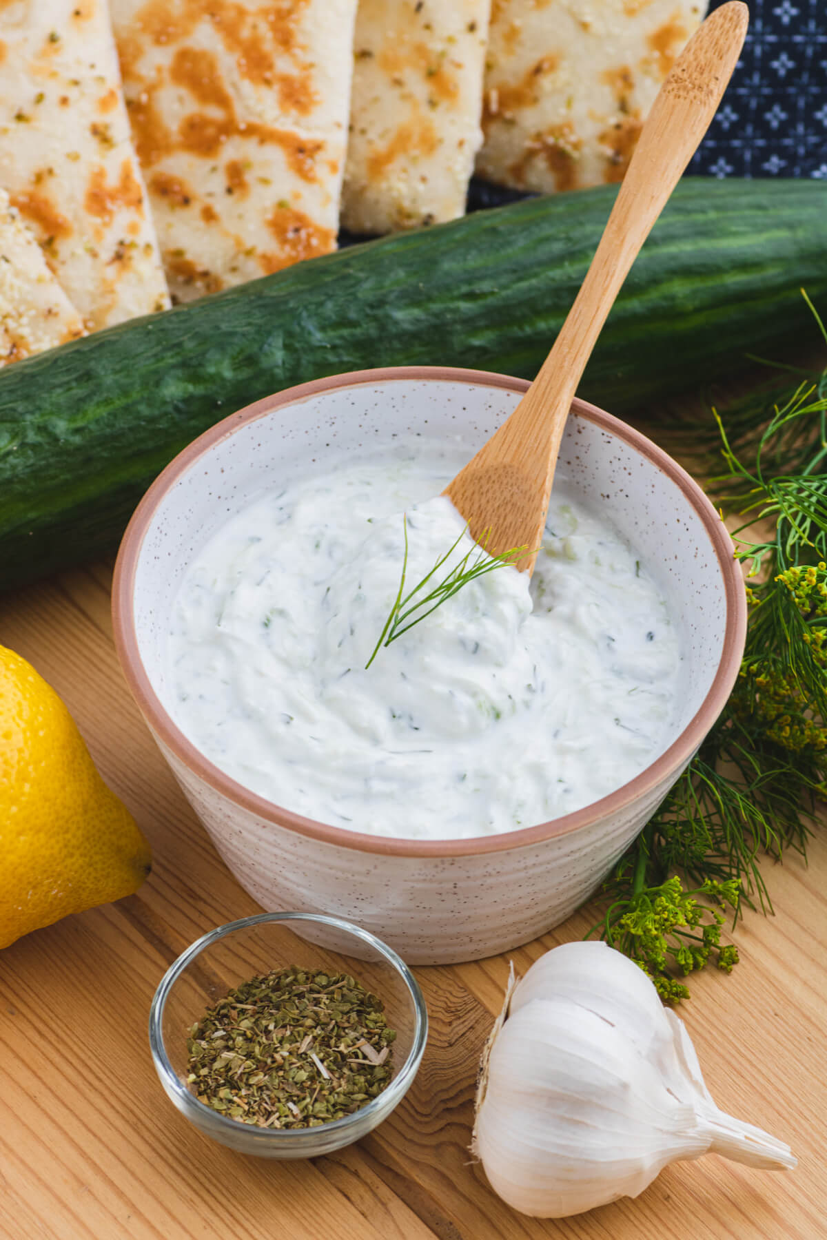 A wooden spoon serves up creamy white Tzatziki sauce flecked with cucumber and dill from a small bowl surrounded by grilled pita bread and other ingredients.