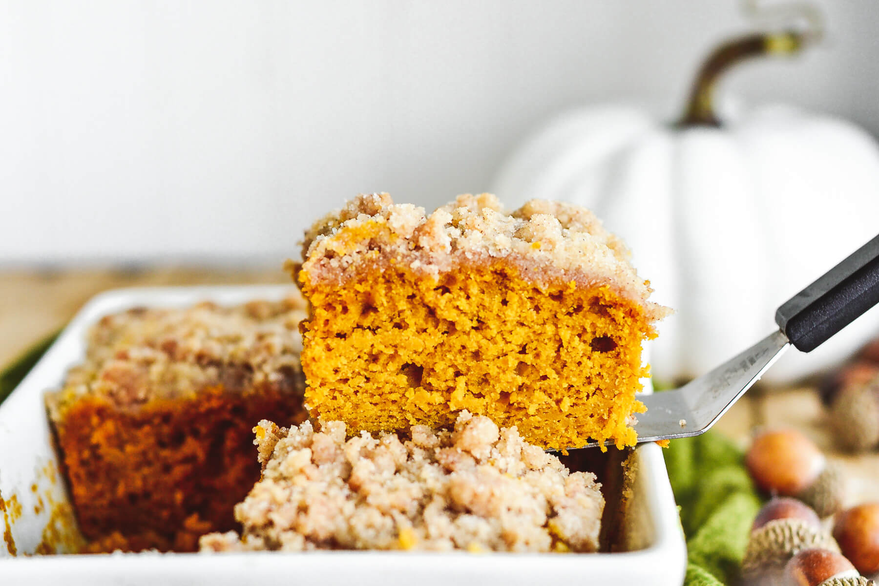 A square of fluffy orange pumpkin coffee cake topped with streusel being removed from a white baking dish.