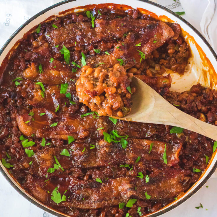 A wooden spoon holding baked beans with ground beef above a skillet filled with the same baked beans.