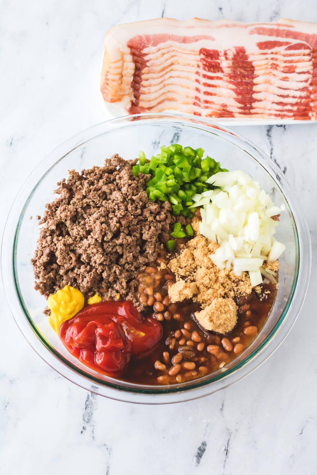A glass mixing bowl filled with ingredients required to make baked beans with ground beef before they are mixed together.