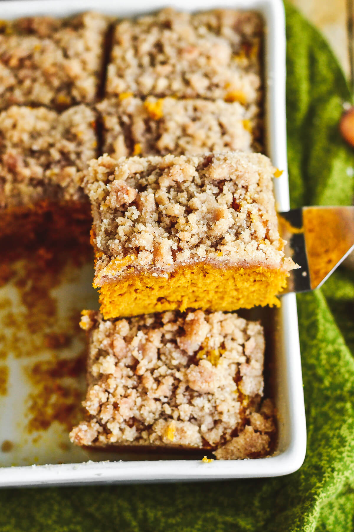 A square of fluffy orange pumpkin coffee cake topped with streusel being removed from a white baking dish.