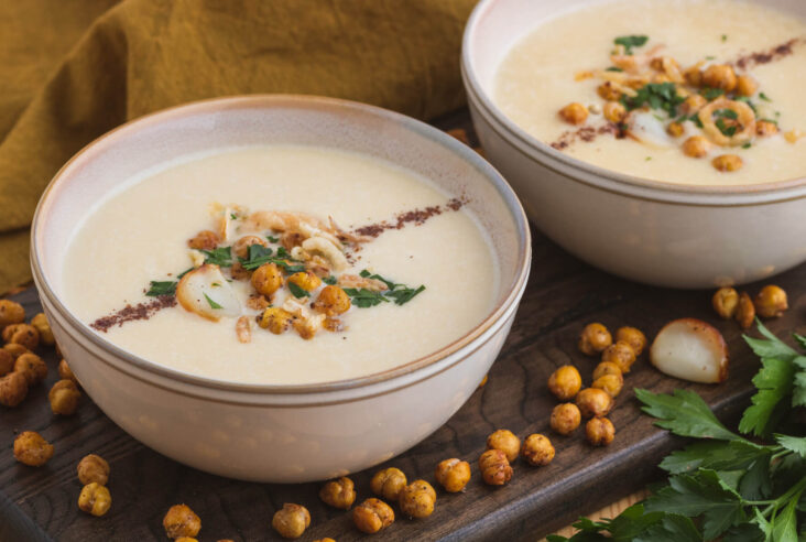 A bowl of creamy Chickpea Soup garnished with sumac, roasted garlic, crispy chickpeas, fried onions, and parsley.
