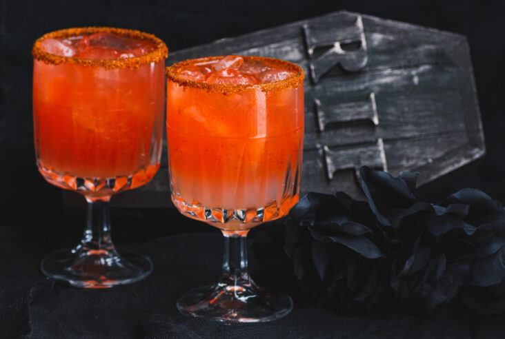 Two blood red Vampiro Cocktails in a Tajin rimmed glass in front of a spooky casket.