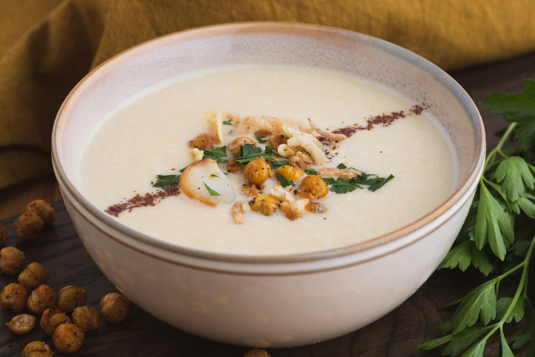 A bowl of creamy Chickpea Soup garnished with sumac, roasted garlic, crispy chickpeas, fried onions, and parsley.