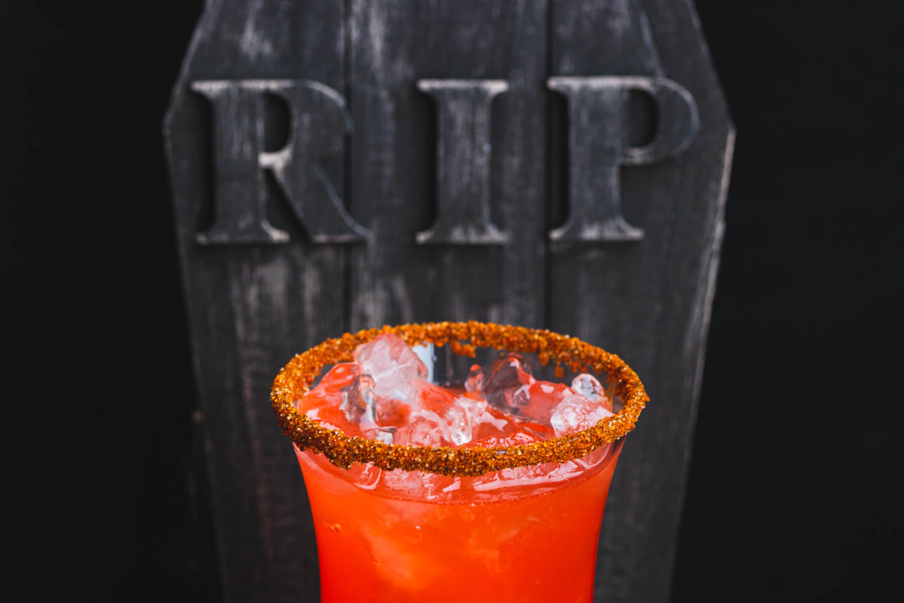 Close up of the Tajin rim on a hurricane glass containing a layered blood red Vampiro Cocktail in front of a creepy tombstone.