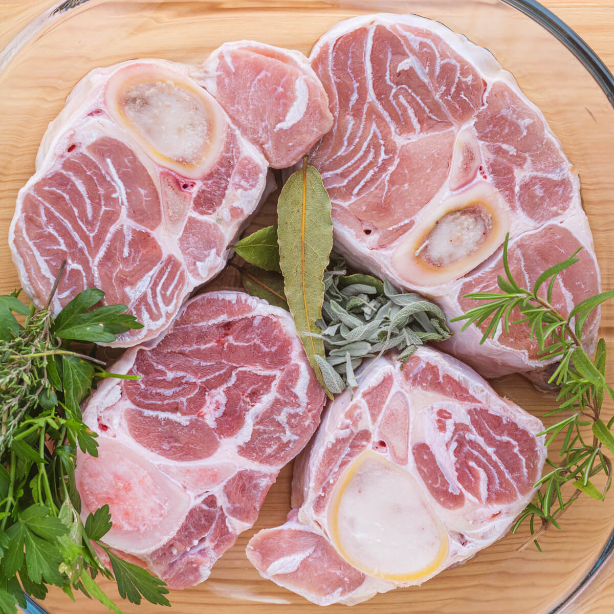 Four raw veal osso bucco surrounded by aromatic green herbs.