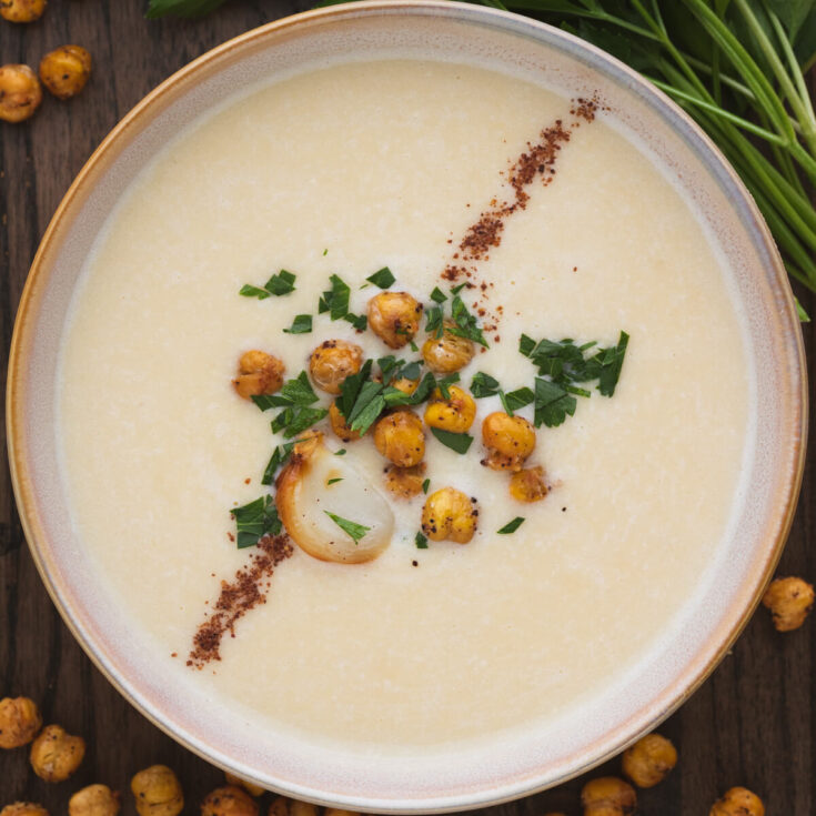 A bowl of creamy Chickpea Soup garnished with sumac, roasted garlic, crispy chickpeas, and parsley.