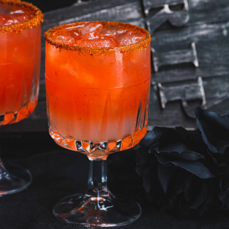Two blood red Vampiro Cocktails in a Tajin rimmed glass in front of a spooky casket.