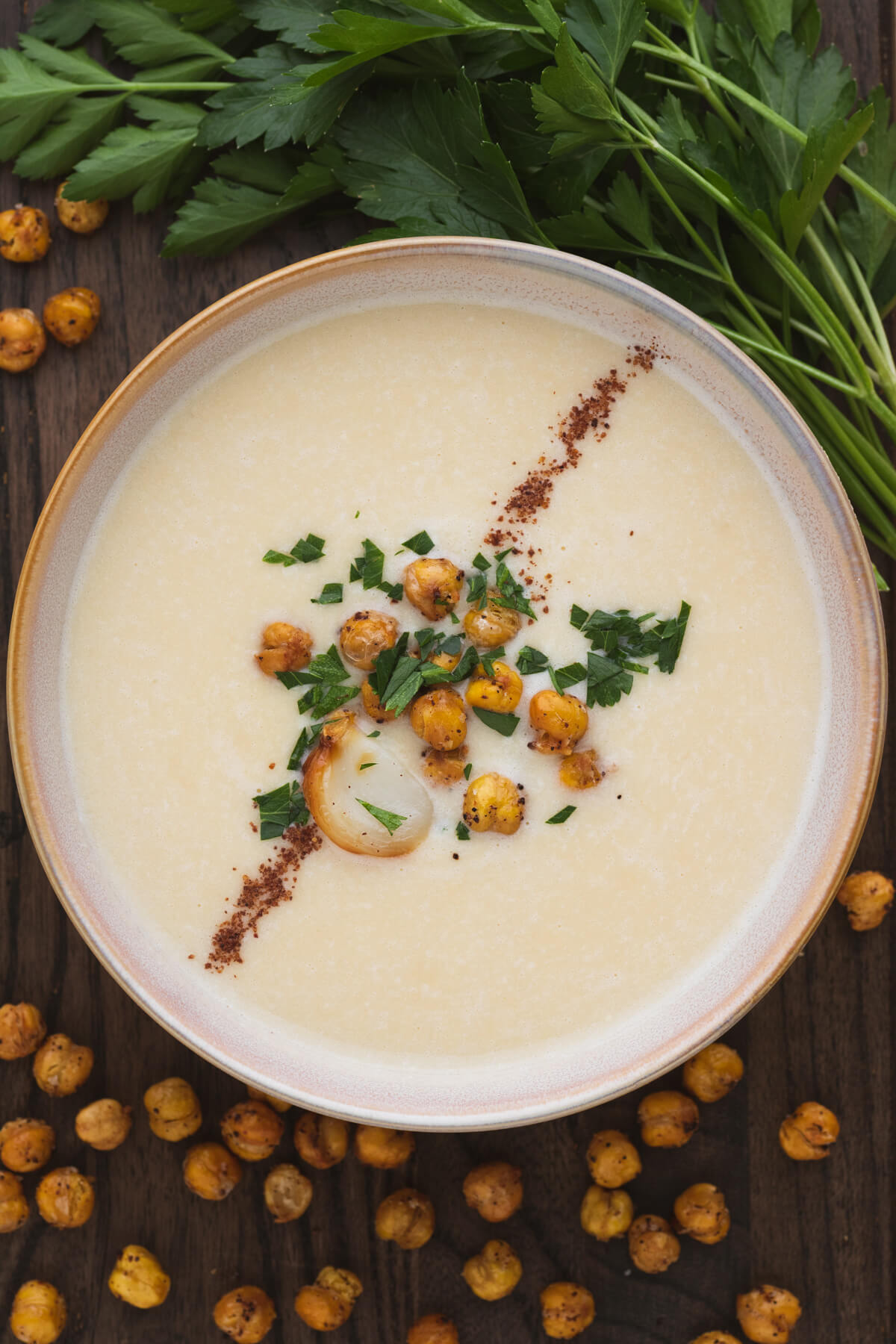 A bowl of creamy Chickpea Soup garnished with sumac, roasted garlic, crispy chickpeas, and parsley.