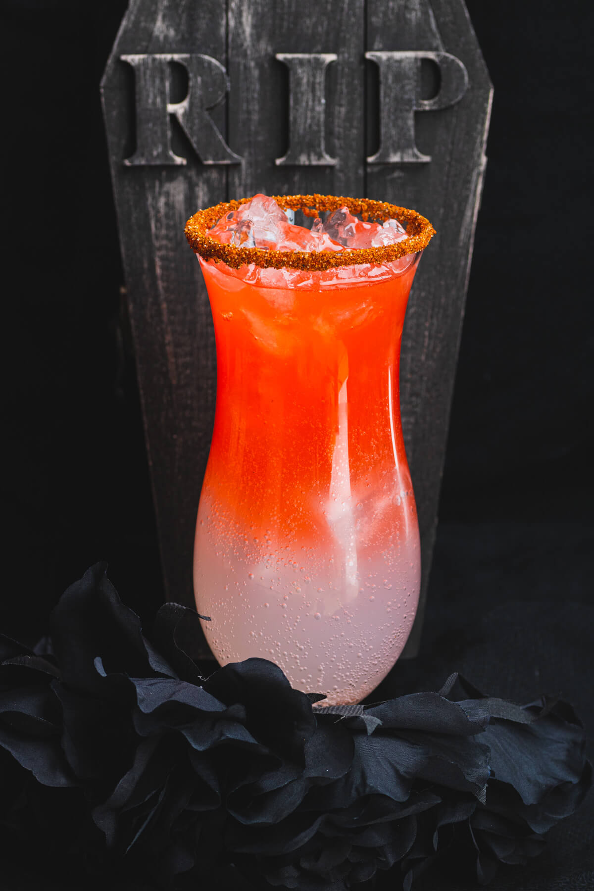 A hurricane glass containing a layered blood red Vampiro Cocktail in front of a creepy tombstone.