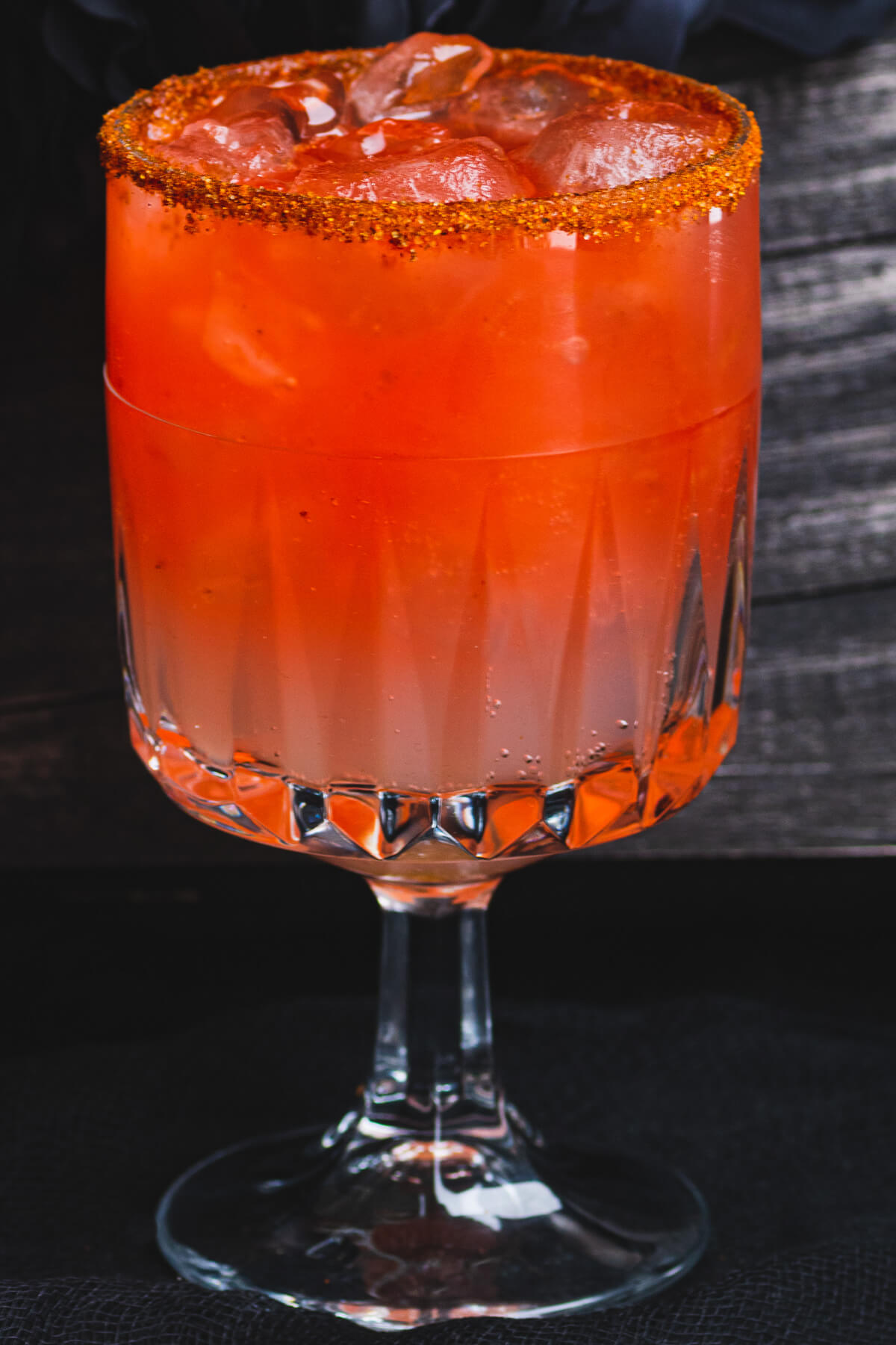 A blood red Vampiro Cocktail in a Tajin rimmed glass in front of a spooky casket.