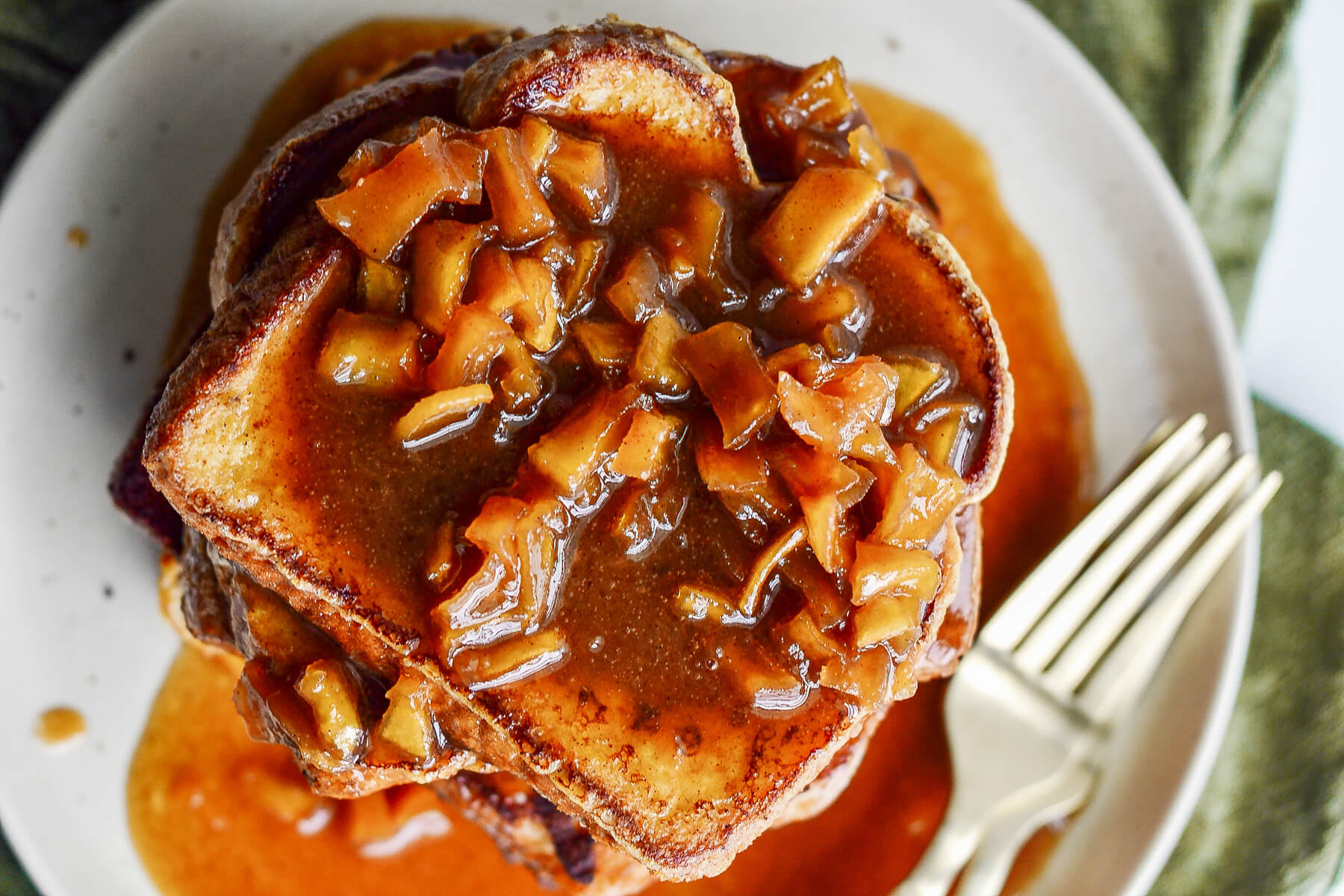 A stack of golden apple French toast topped with a rich caramel sauce on a white plate.