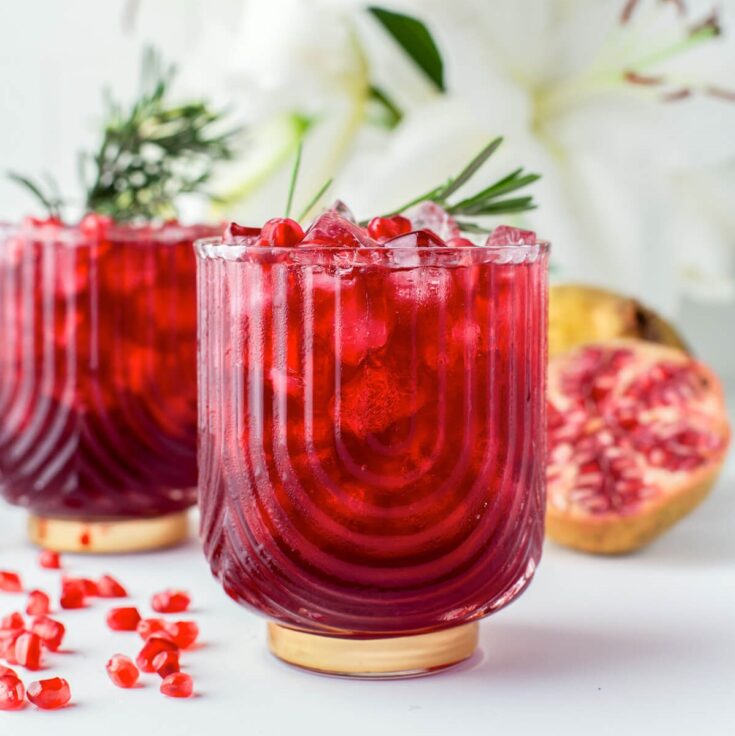 Two stunning ruby red cocktails in rocks glasses garnished with pomegranate arils and a sprig of rosemary.