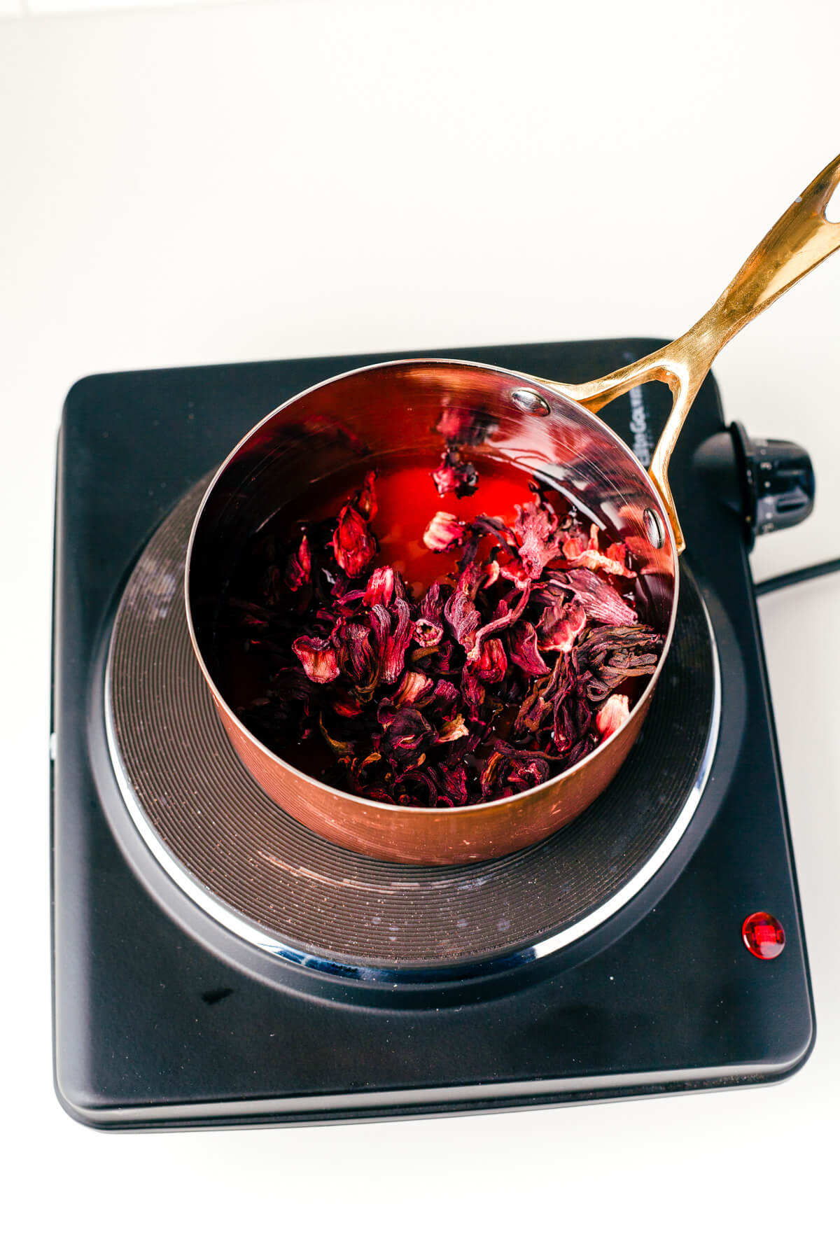 Showing how to make cocktail syrup by infusing pomegranate juice with hibiscus and sugar in a saucepan.