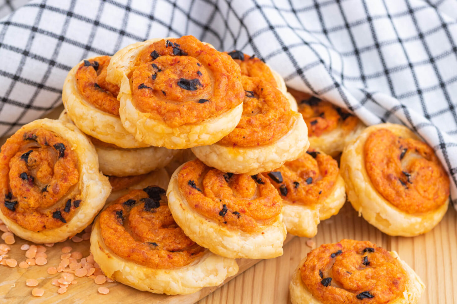 A pile of golden baked puff pastry pinwheels on a wooden board garnished with red lentils.