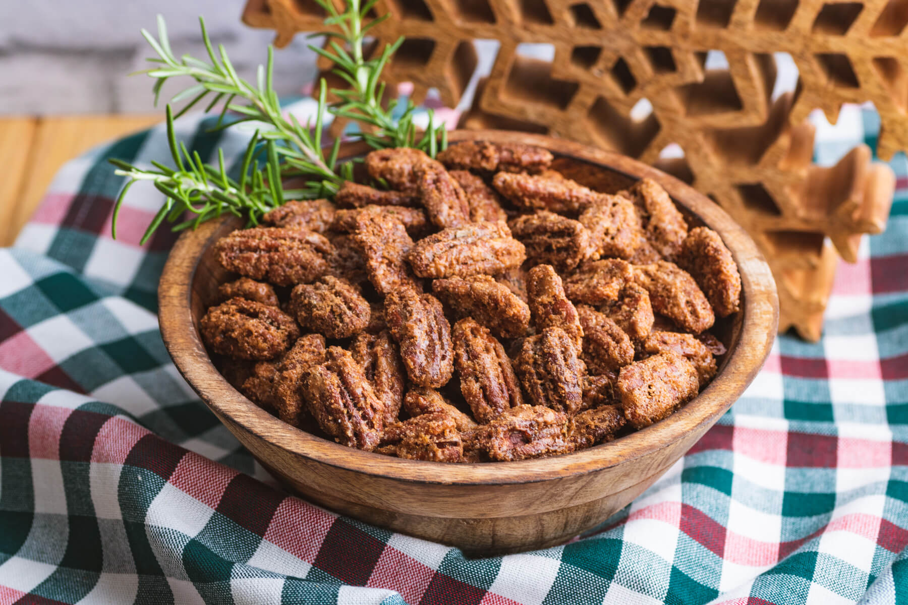A wooden bowl filled with candied pecan halves and fresh rosemary garnish on a plaid tablecloth.