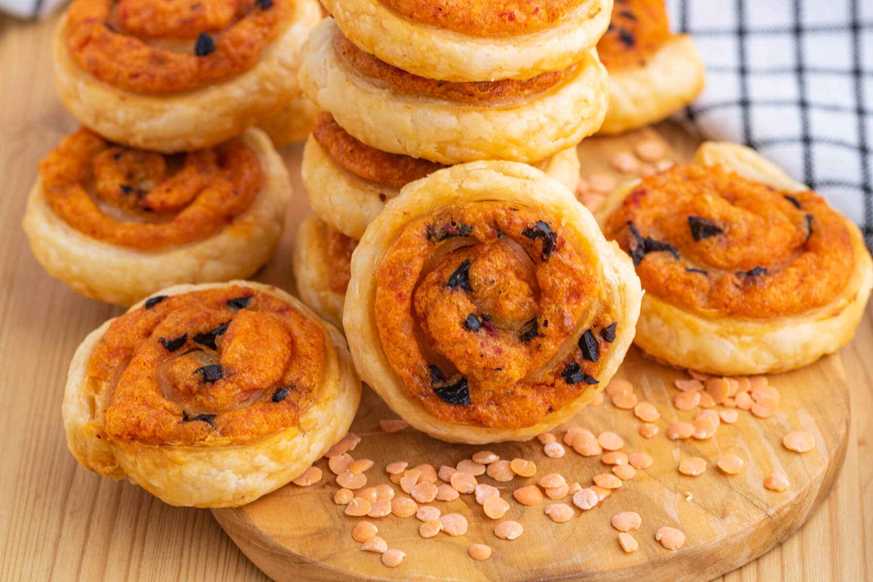 A pile of golden baked puff pastry pinwheels on a wooden board garnished with red lentils.
