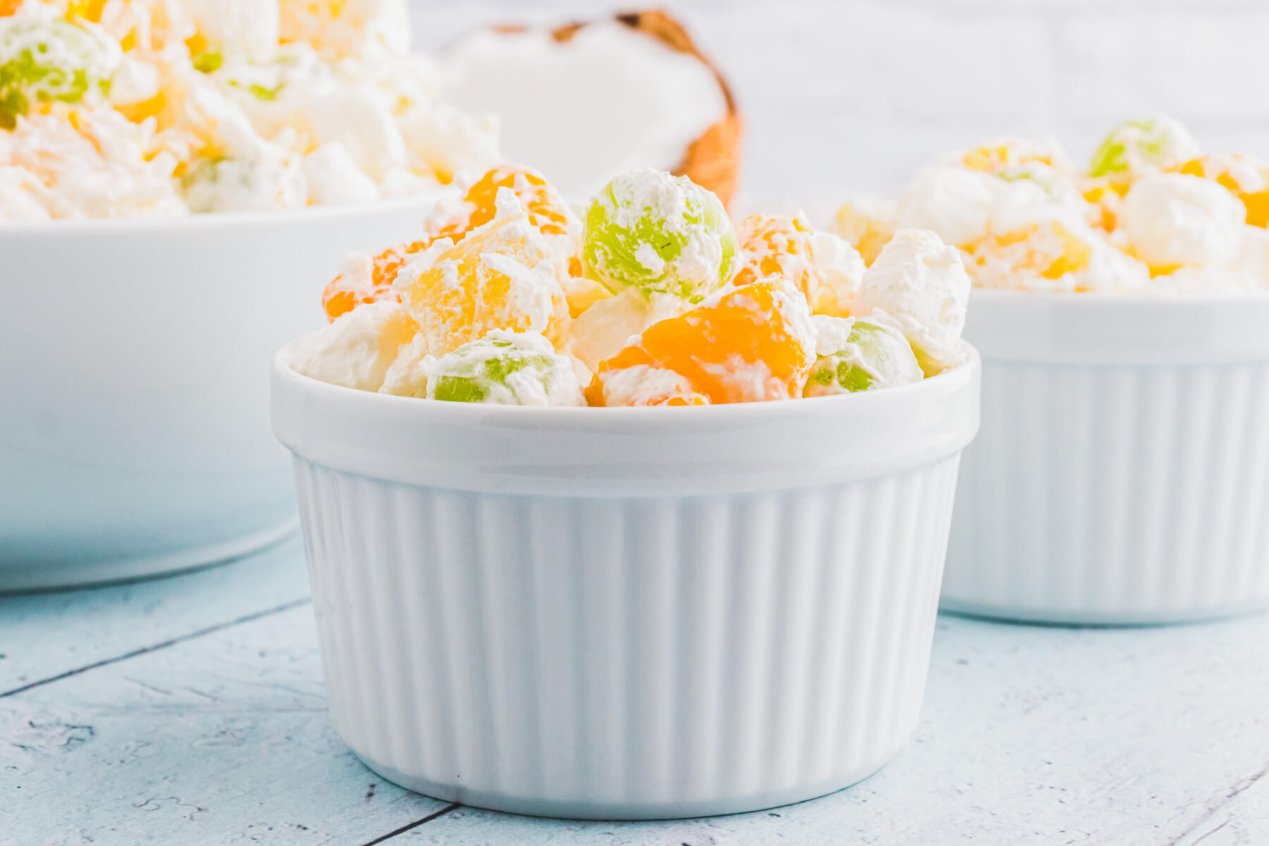 Three white bowls of fluffy dreamy Ambrosia fruit salad on a light blue background.