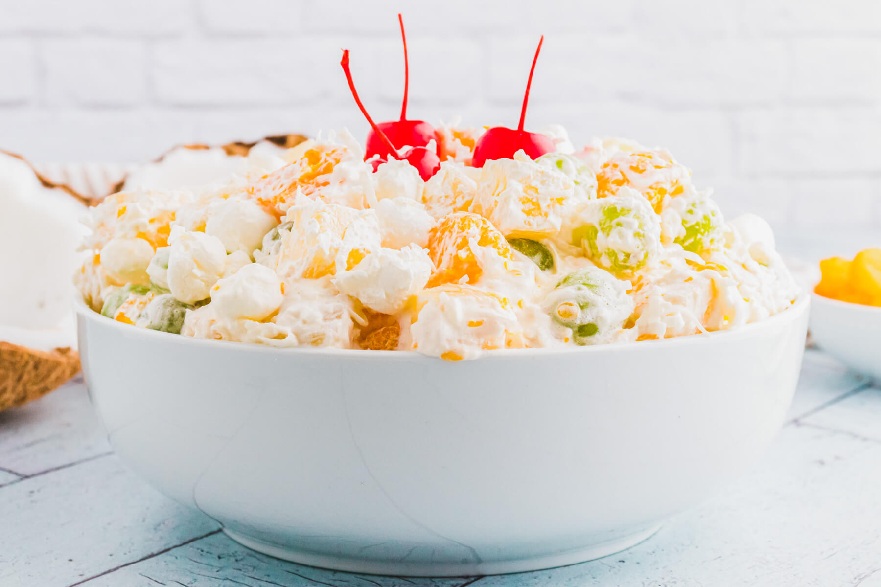 A white bowl of fluffy dreamy Ambrosia fruit salad garnished with maraschino cherries on a light blue background.
