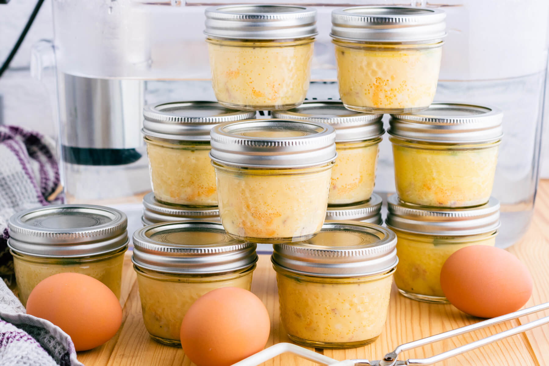Stacks of Sous Vide Egg Bites in glass jars and brown eggs in front of a sous vide container.