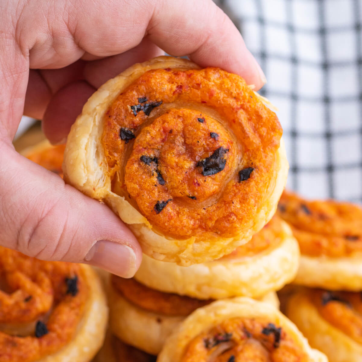 A hand holds one puff pastry pinwheel above a pile of the same appetizers.