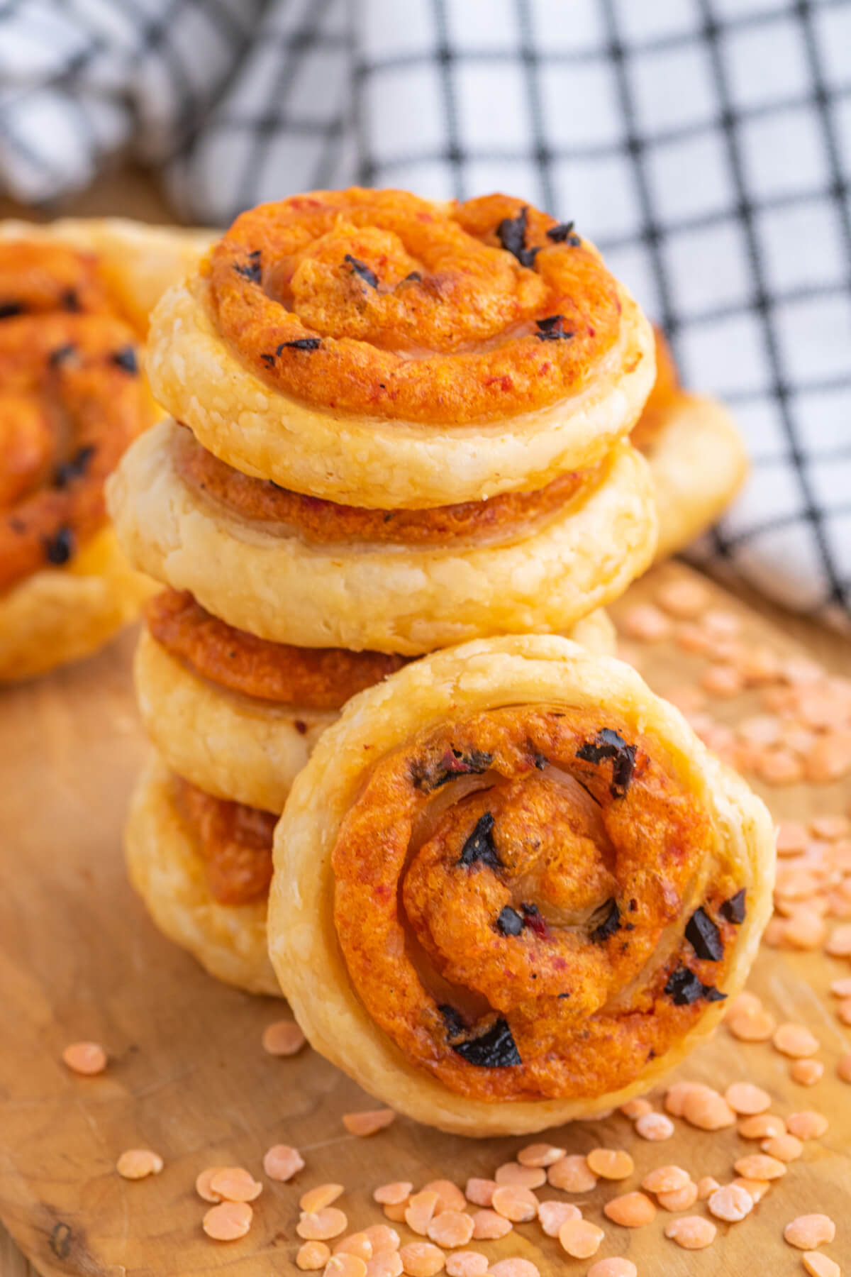 A stack of golden baked puff pastry pinwheels on a wooden board garnished with red lentils.