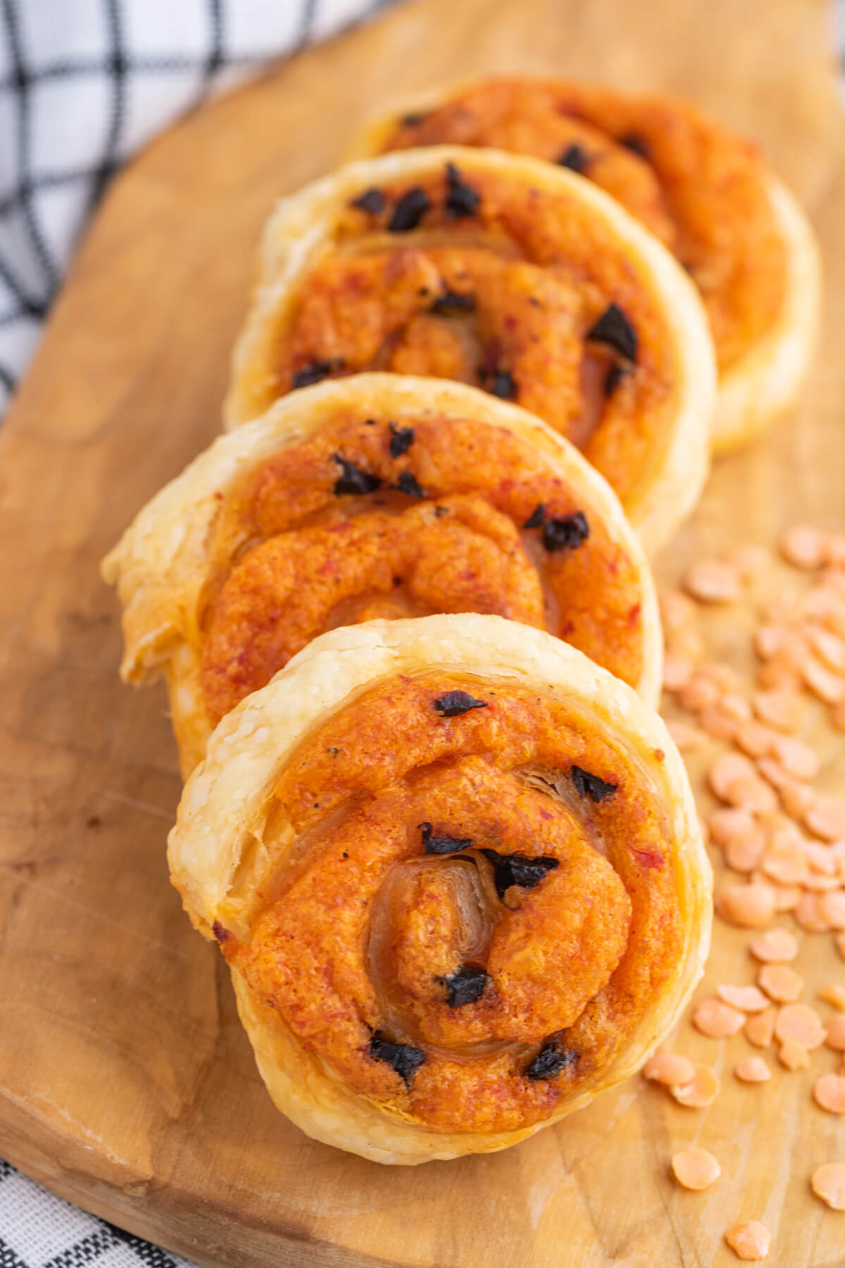 A row of golden baked puff pastry pinwheels on a wooden board garnished with red lentils.