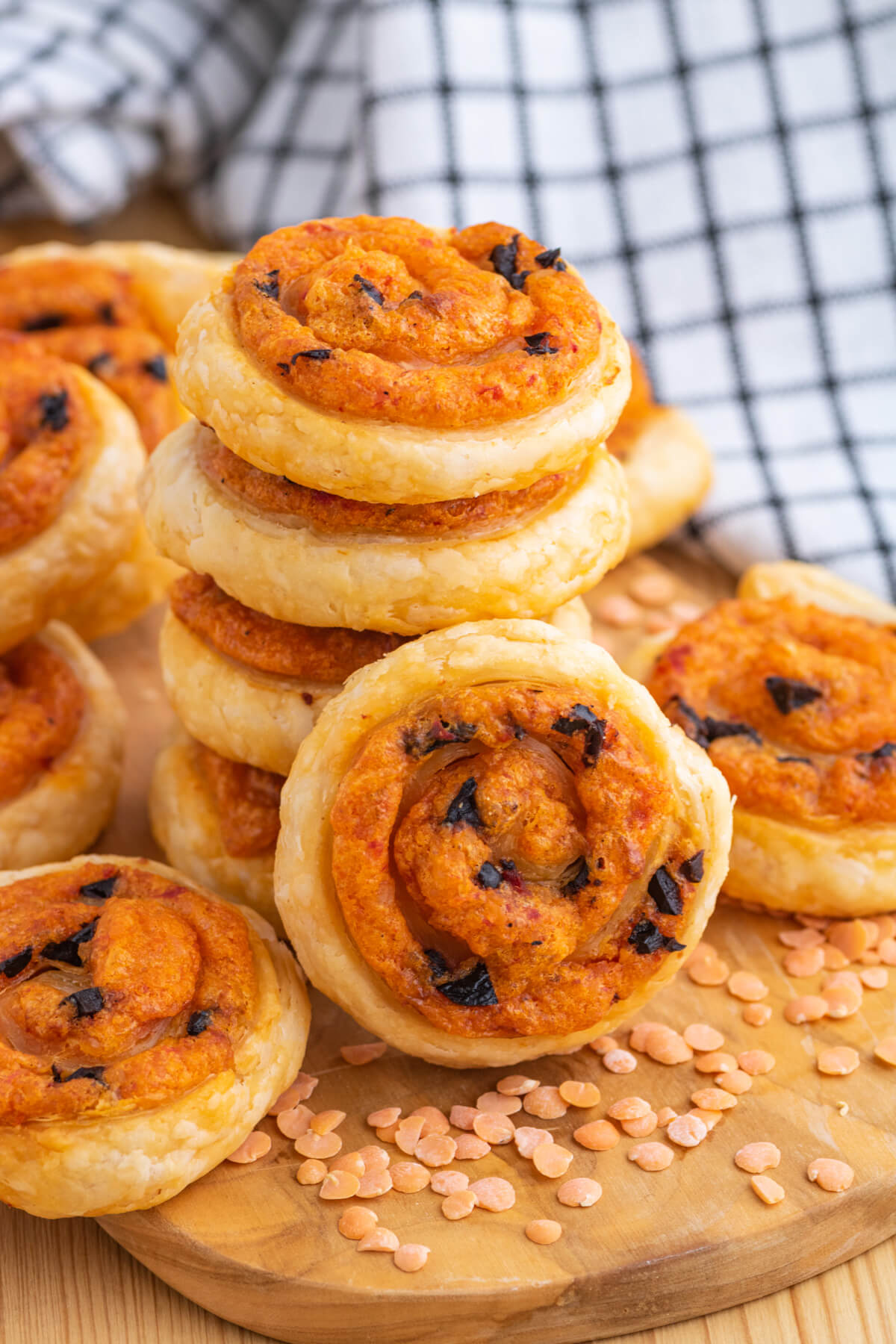 A stack of golden baked puff pastry pinwheels on a wooden board garnished with red lentils.