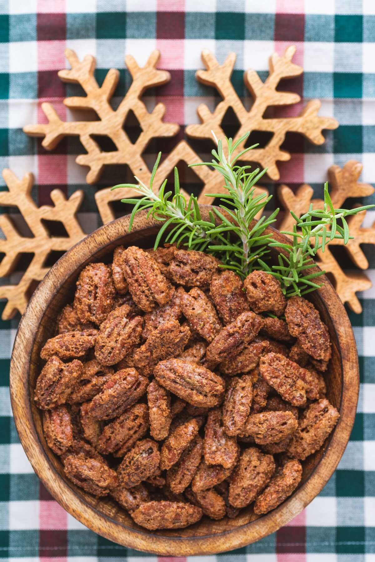 A wooden bowl filled with candied pecan halves and fresh rosemary garnish sits on a wooden snowflake trivet.