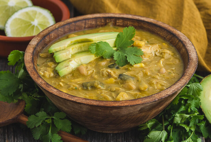 A wooden bowl filled with vibrant green Chicken Chili Verde garnished with cilantro and avocado slices.