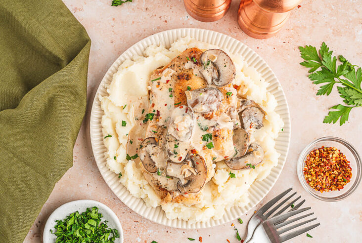A serving of creamy chicken marsala featuring a whole chicken breast topped with mushrooms and creamy sauce over a bed of mashed potatoes.