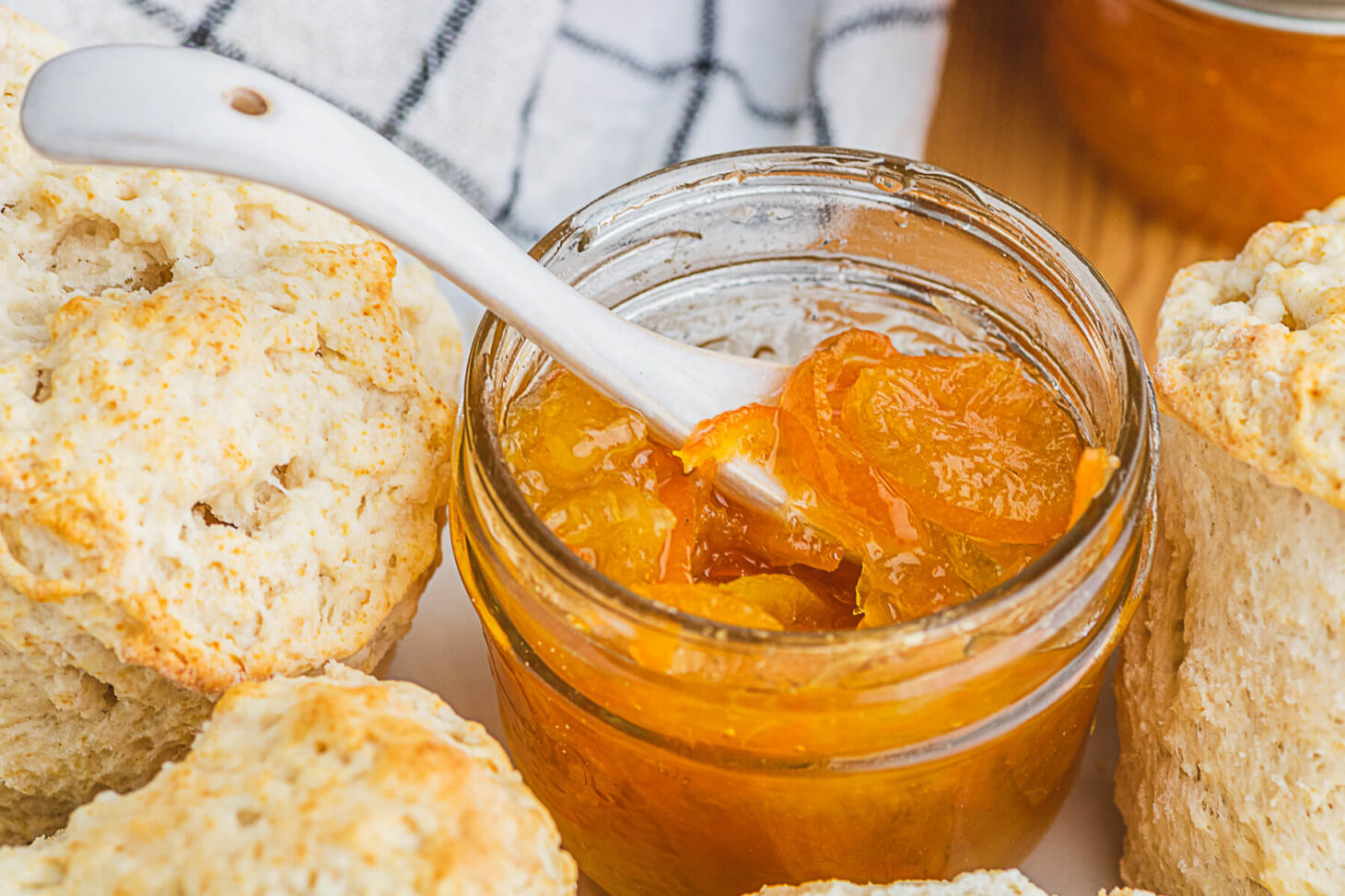 A small jar of bright Kumquat Marmalade with a tiny pottery spoon surrounded by biscuits.