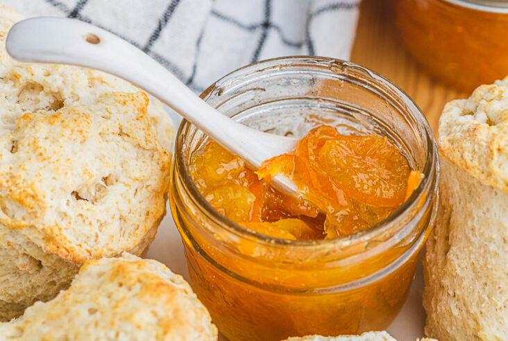 A small jar of bright Kumquat Marmalade with a tiny pottery spoon surrounded by biscuits.