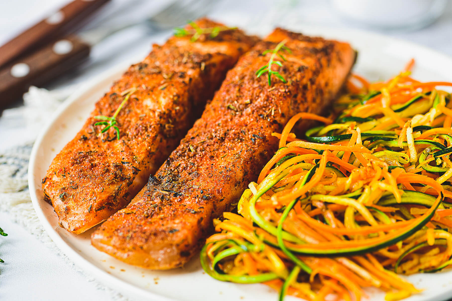 Two cooked spiced salmon fillets on a plate beside spiralized vegetables.