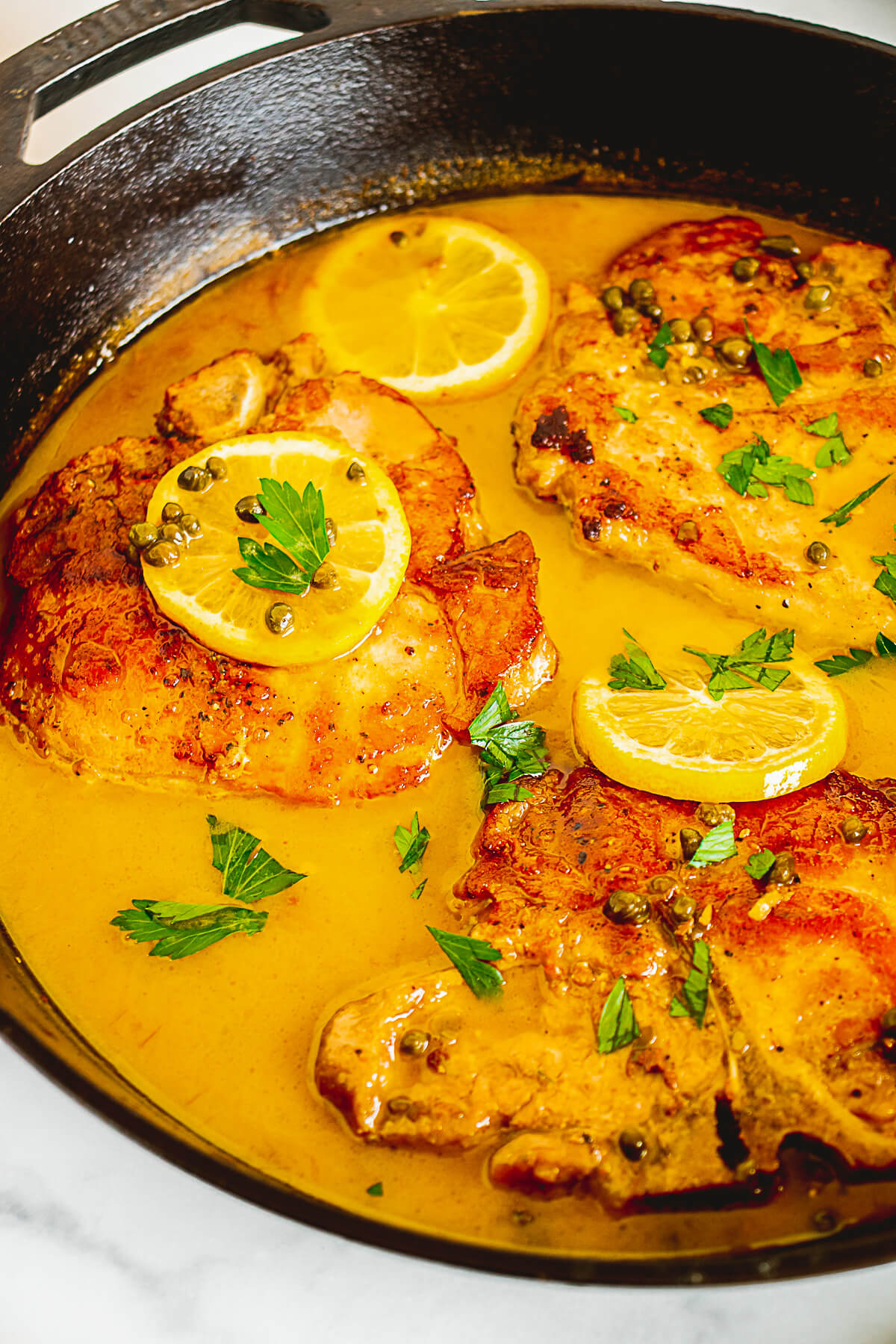 Three golden brown pork chops in a cast iron skillet coated in a luxurious lemony piccata sauce garnished with lemon slices and fresh parsley.
