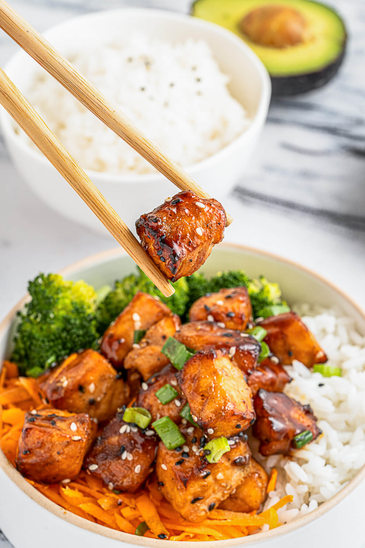 A pair of wooden chopsticks hold a piece of teriyaki salmon over a bowl of teriyaki salmon, vegetables, and rice.