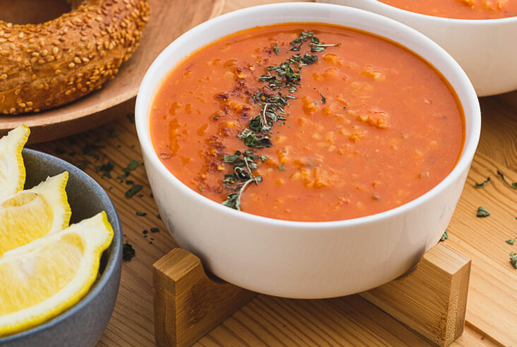 A white bowl filled with fiery red Turkish Lentil Soup garnished with dried mint and red pepper flakes surrounded by lemon wedges and bread.